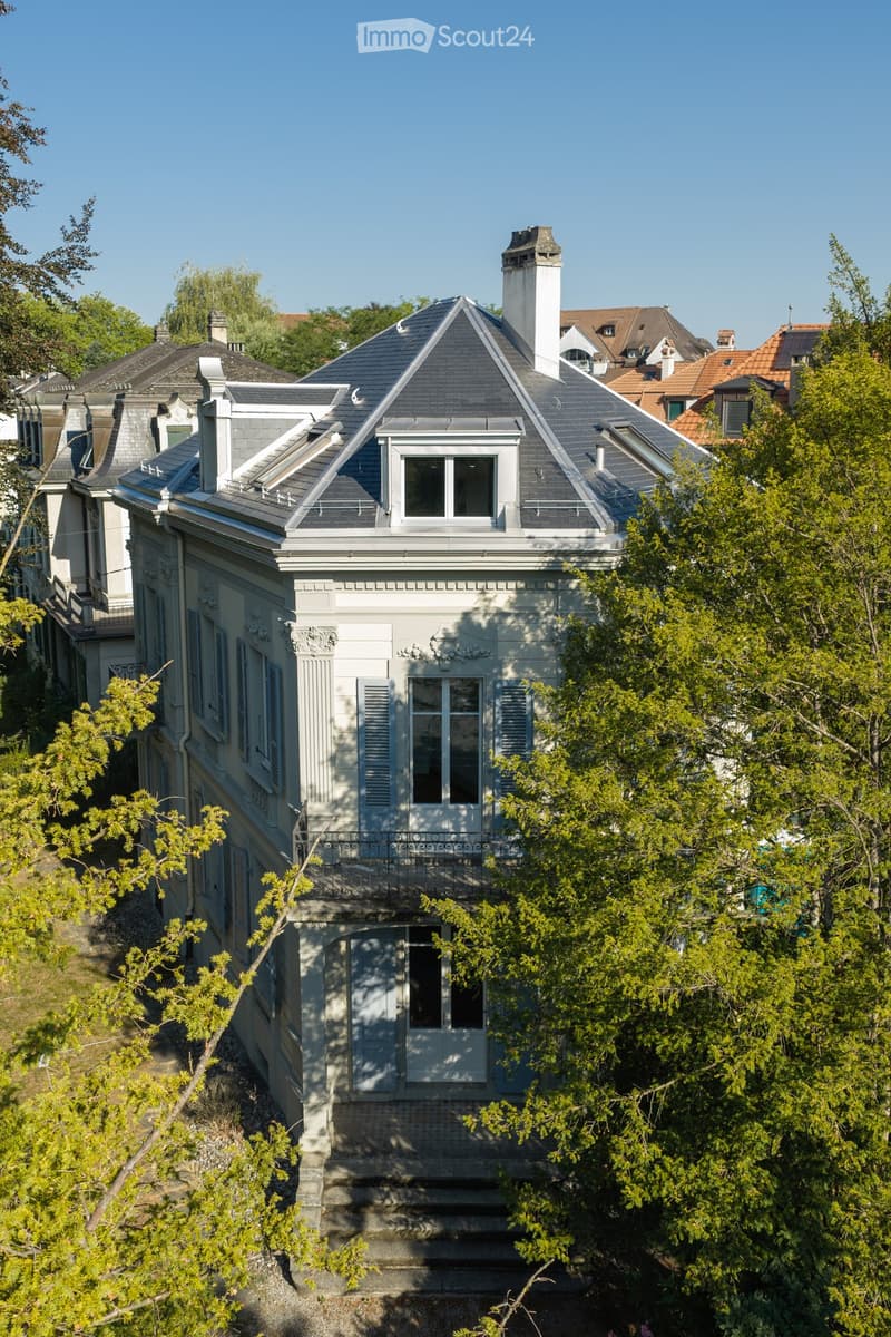 A multi-story house with a grey roof, white walls, and ornate architectural details, surrounded by trees and greenery.