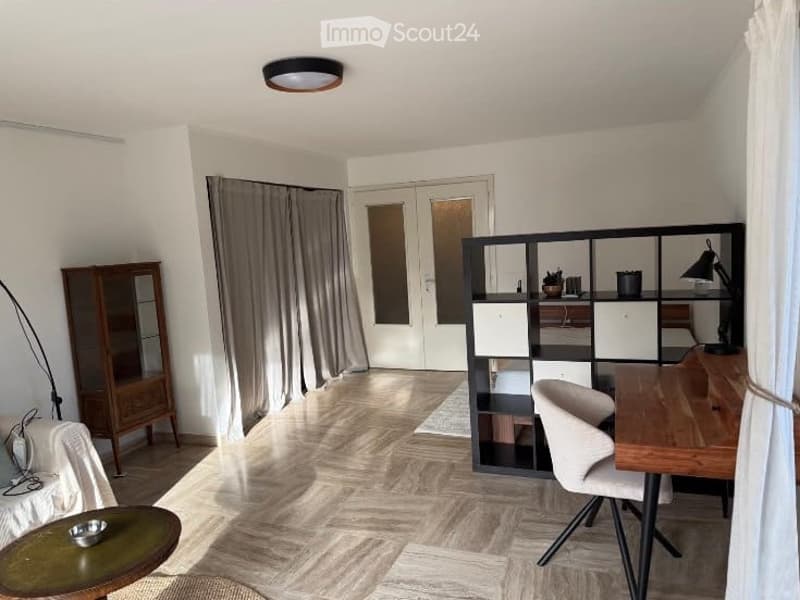Bright, modern living space with a mix of living and working areas. Features a sofa, coffee table, wooden cabinet, large shelving unit, desk with chair, and floor lamp. Natural light streams in through large windows with curtains.