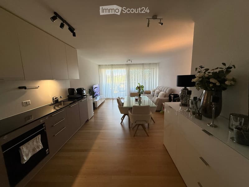 Open-plan living and dining area with modern kitchen, light wood flooring, white cabinets, and large windows with curtains. Features a sofa, coffee table, dining table with chairs, and decorative vases.