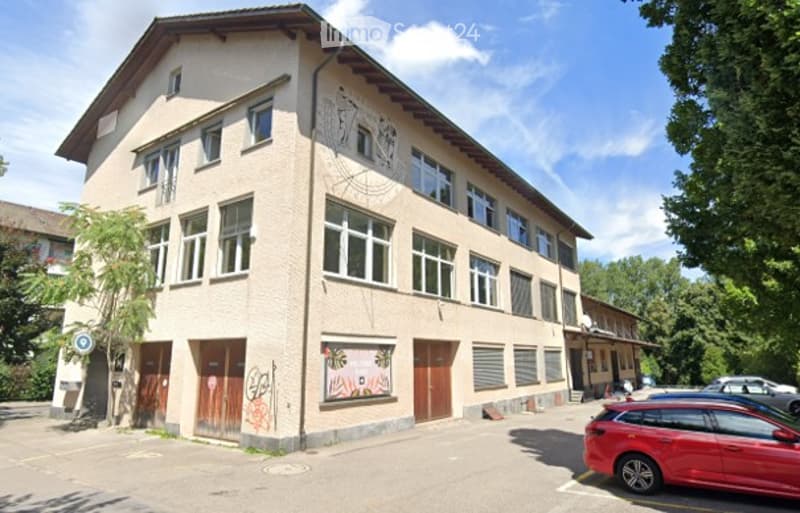 Two-story building with beige facade, multiple windows, and a mural on the side. There is a parking area with a red car in front.
