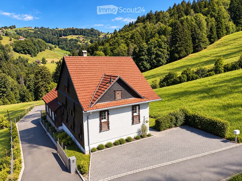 A two-story house with a red-tiled roof and white walls, situated in a green, hilly landscape with trees and open fields.