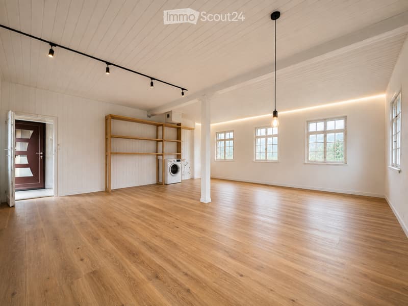 Bright, airy room with wooden flooring, white paneled walls, and exposed wooden beams on the ceiling. Features built-in wooden shelving and a washing machine in the corner, with large windows allowing natural light to fill the space.