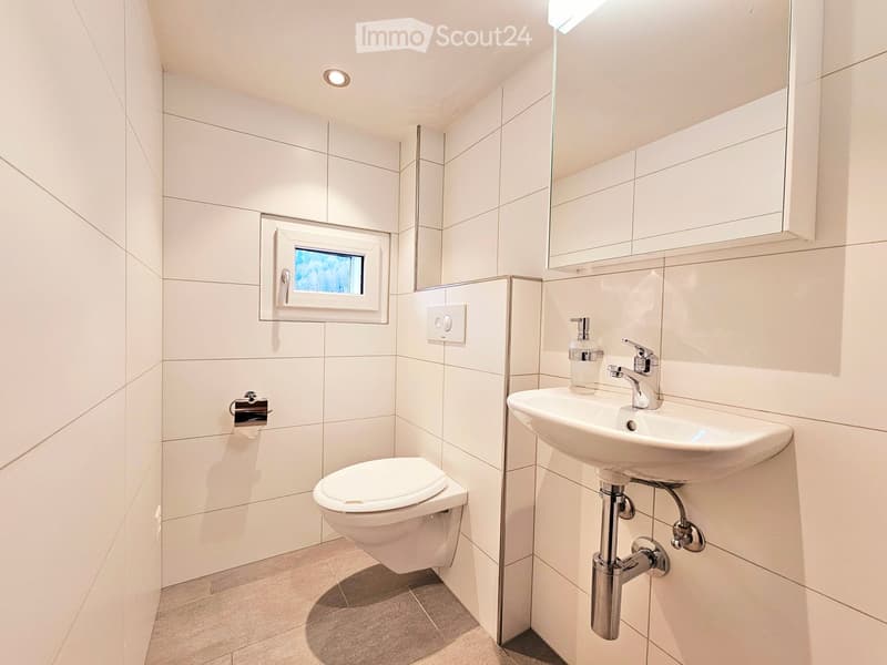 Modern bathroom with white tiles, a toilet, a sink with a modern faucet, a mirror, and a small window providing natural light.