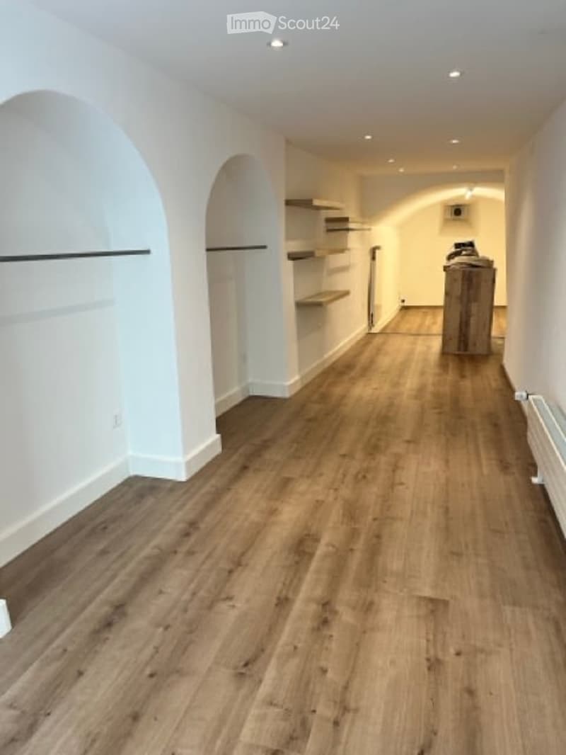 Empty room with hardwood floors, white walls, arched doorways, wooden shelves, power outlet, wooden stool, radiator.
