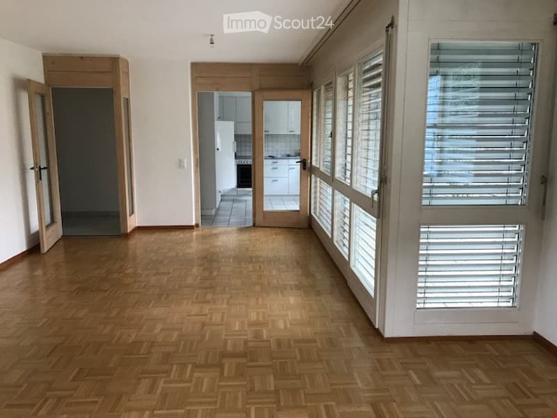 Empty room with wooden parquet flooring, white walls, large windows with blinds, and open doors leading to a kitchen area.