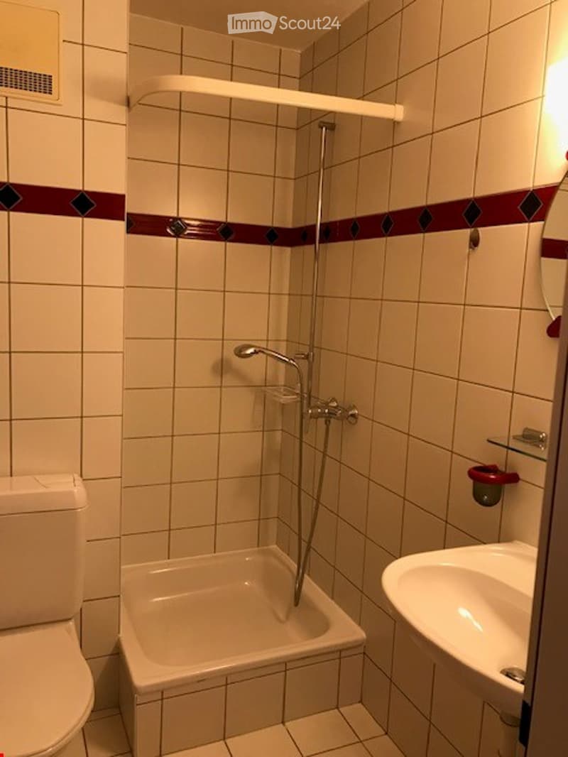 A compact bathroom with white tiled walls and floor, featuring a toilet, a small shower stall, a sink with a mirror, and a shelf above the shower.