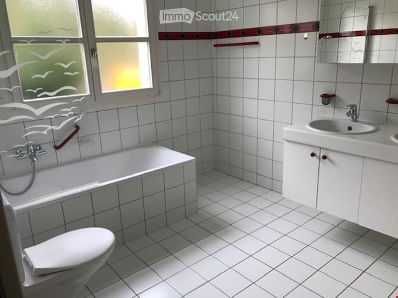 A clean, white-tiled bathroom with a bathtub, toilet, sink, and mirror. The window has a decorative bird design on the glass.