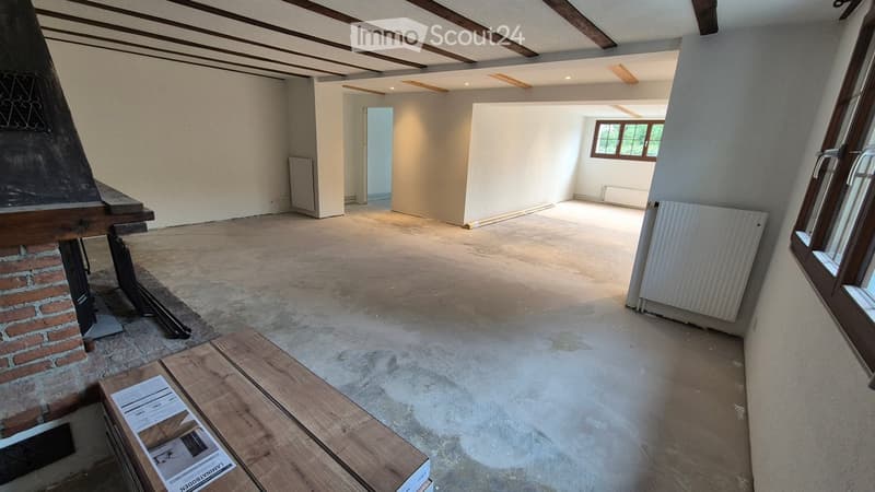 Empty room with exposed wooden beams on the ceiling, concrete floor, and a brick fireplace on the left side. There are no furnishings or decorations visible.