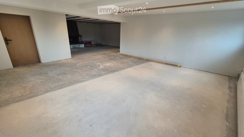 Empty room with bare walls and unfinished concrete floor, featuring a wooden door and a small open area with a fireplace and some stacked items.