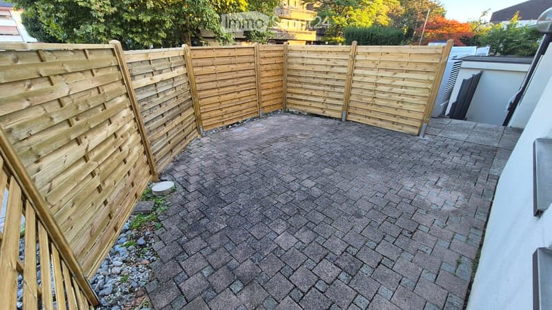 A paved outdoor area enclosed by a wooden fence, with a mix of brick and stone tiles, small patches of grass, and some rocks near the fence.