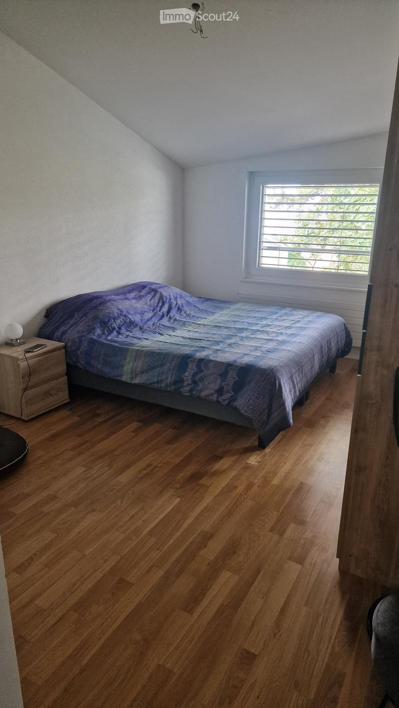 Cozy bedroom with wooden flooring, a single bed with a patterned blue and white duvet, a nightstand with a lamp and a cellphone, and a window with blinds.