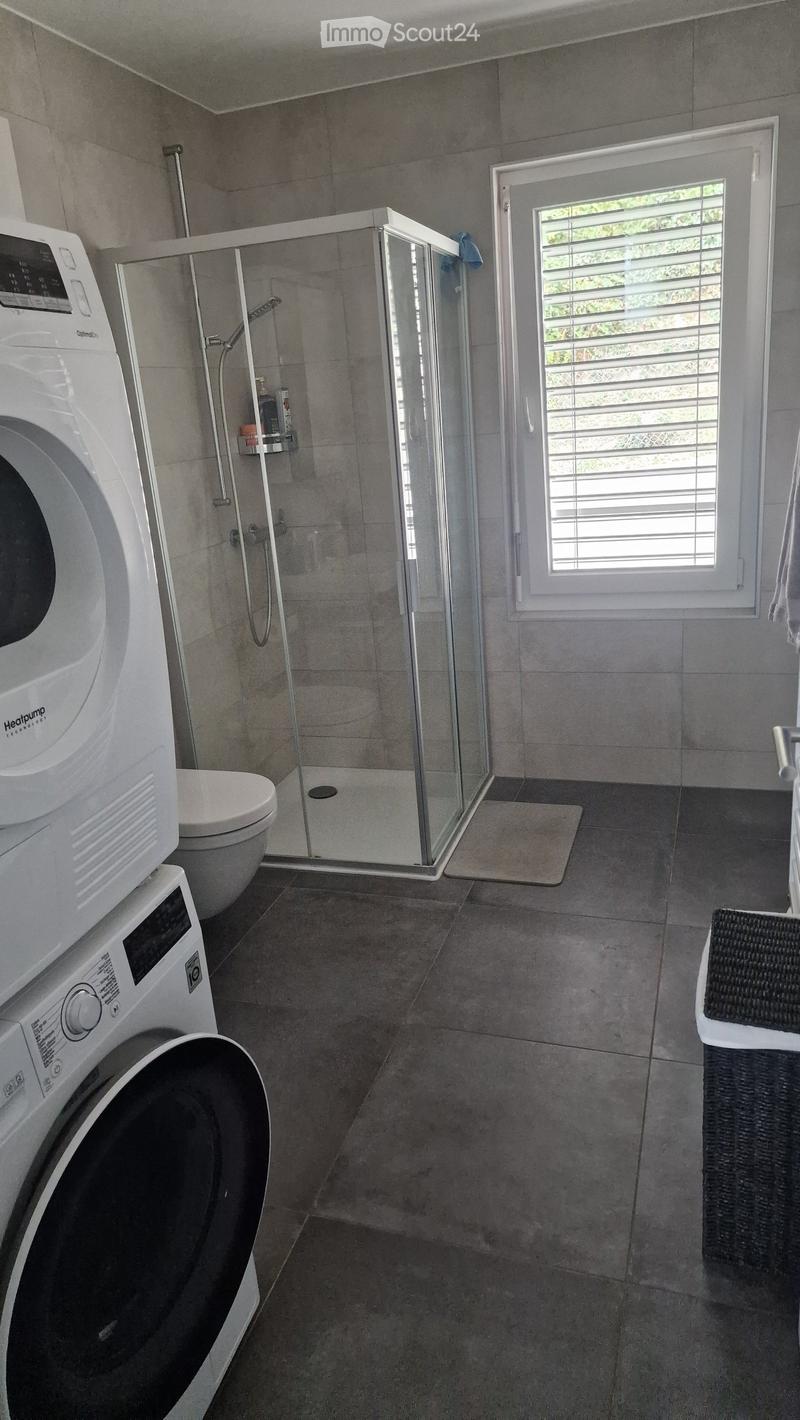 Modern bathroom with a glass-enclosed shower, toilet, washing machine, and dishwasher. Features tiled walls and floor, a window with blinds, and a small shelf inside the shower.