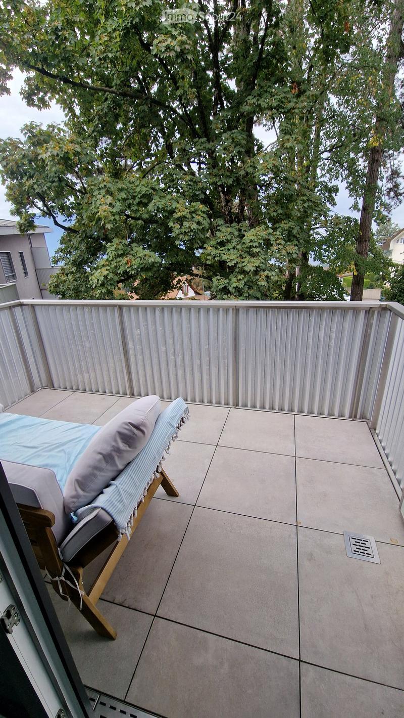 Balcony with a sun lounger, light blue cover, and grey cushions, surrounded by trees and a metal railing.