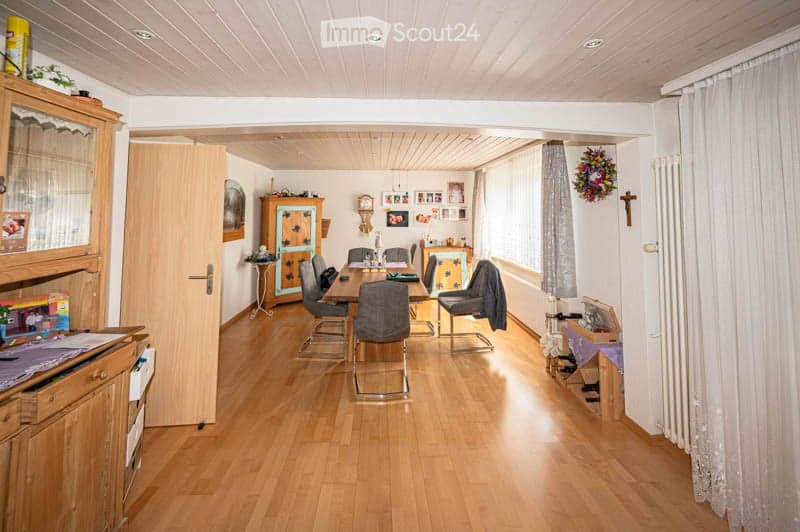 Bright dining room with wooden flooring, a dining table surrounded by chairs, a wooden cupboard, and a radiator near the window with curtains.