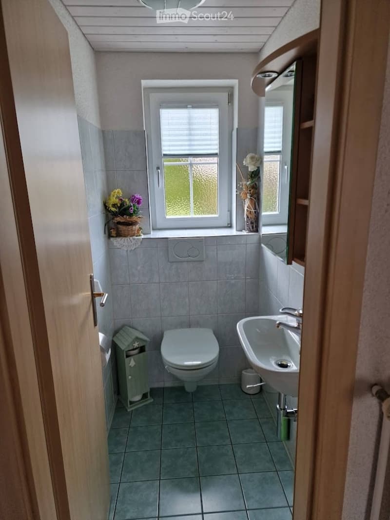 Cozy bathroom with a toilet, sink, mirror, and decorative flower vase on the windowsill. The room features light grey tiles and a small window with blinds.