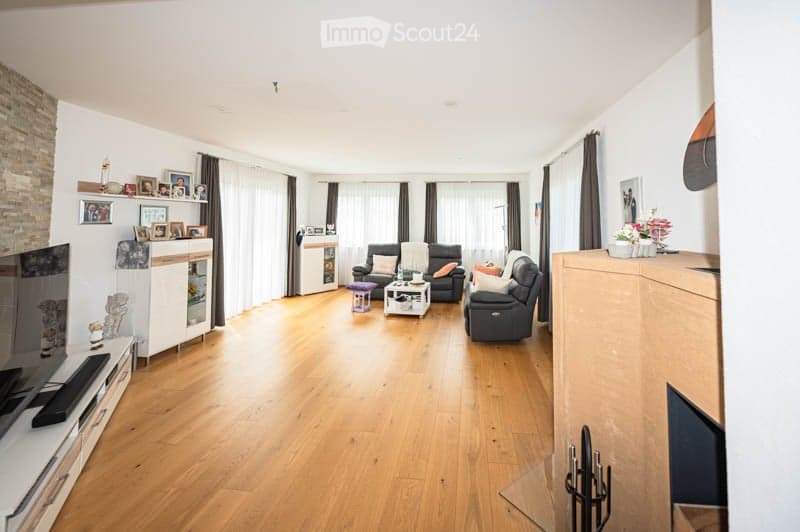 Bright living room with hardwood flooring, white walls, large windows with curtains, and a mix of modern and classic furniture including a sofa, coffee table, TV stand, and decorative items on shelves.