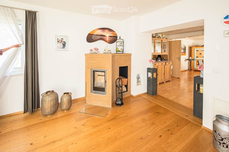 A modern living room with a central fireplace, wooden flooring, and large windows. The space includes a cabinet, a small table with a vase, and decorative items such as picture frames and vases. The room is well-lit and has a cozy, inviting atmosphere.