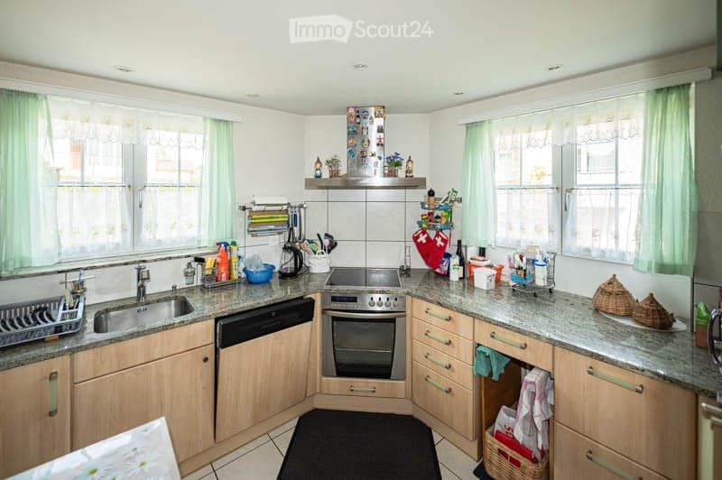 The kitchen features light wooden cabinets, granite countertops, and stainless steel appliances including an oven, dishwasher, and sink. The space is well-lit with natural light from windows, and various kitchen items such as a toaster, cups, and cleaning