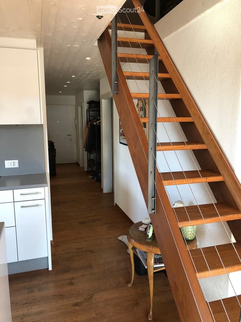A modern hallway with wooden flooring, white walls, and a staircase with wooden steps and metal railings. A small round table with a few items sits near the stairs, and a kitchen area is visible on the left side with white cabinets and a countertop.