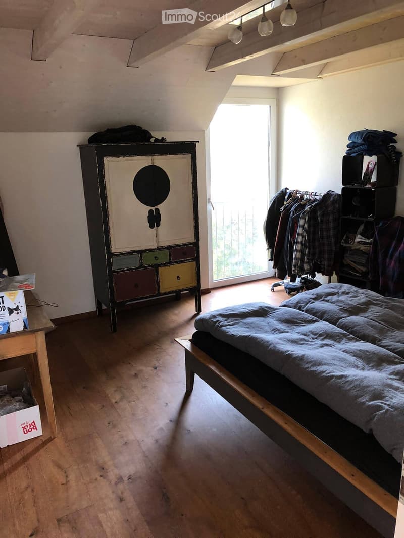 Bedroom with wooden floor, slanted ceiling, bed with blue duvet, wardrobe with colorful drawers, clothing rack, and small table with a box on it.