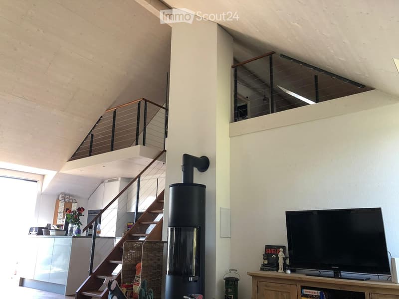 Modern living room with a large television on a wooden stand, a sleek black wood-burning stove, a staircase with a glass railing, and a small kitchen area visible in the background.