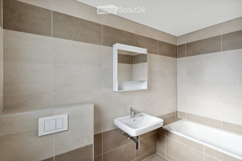 Modern bathroom with beige tiles, a white sink, a bathtub, a wall-mounted mirror, and a light switch.