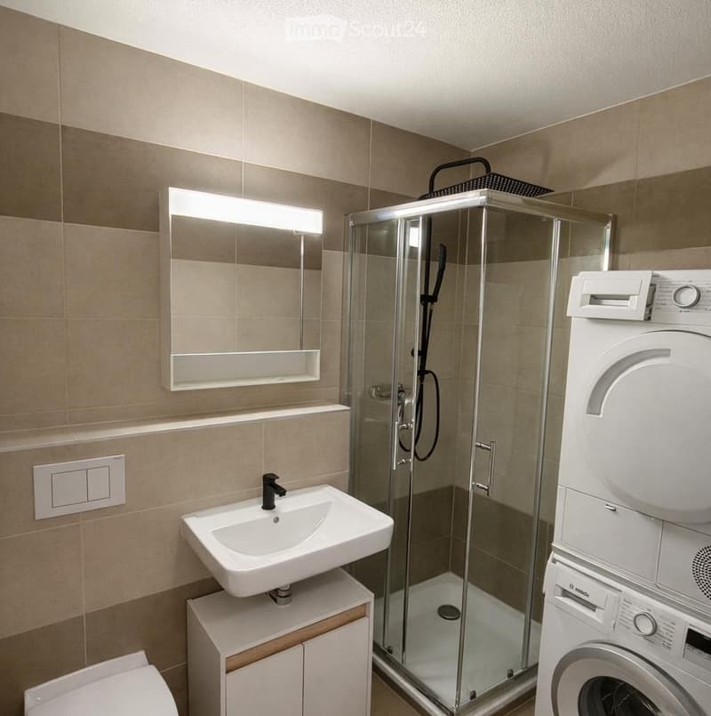Modern bathroom with a white sink, mirror, glass-enclosed shower, toilet, and a stacked washer/dryer unit.