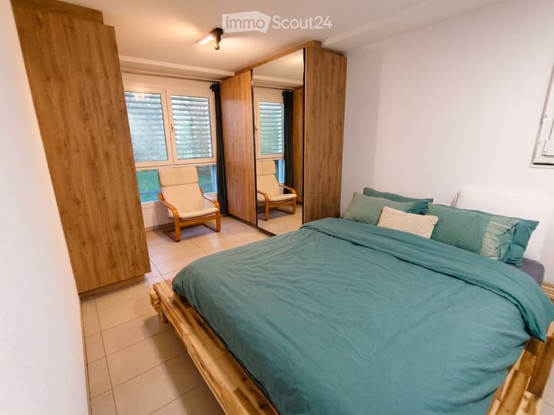 Cozy bedroom with a wooden bed, green bedding, a wooden wardrobe, a mirror, and two wooden chairs near the window.