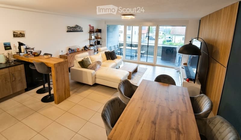 Modern living and dining area with a white sectional sofa, coffee table, dining table with chairs, kitchen counter, and a large balcony with outdoor furniture visible through large windows.