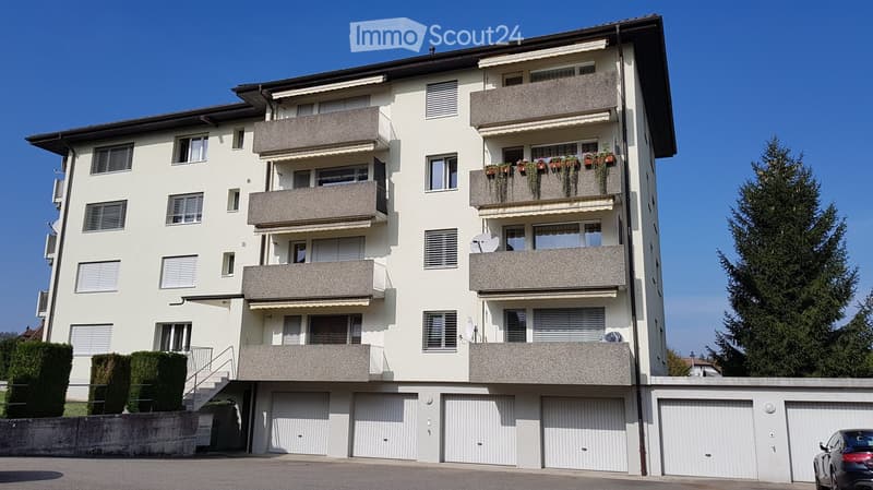 Four-story apartment building with multiple balconies, white exterior, and several garage doors at the base.
