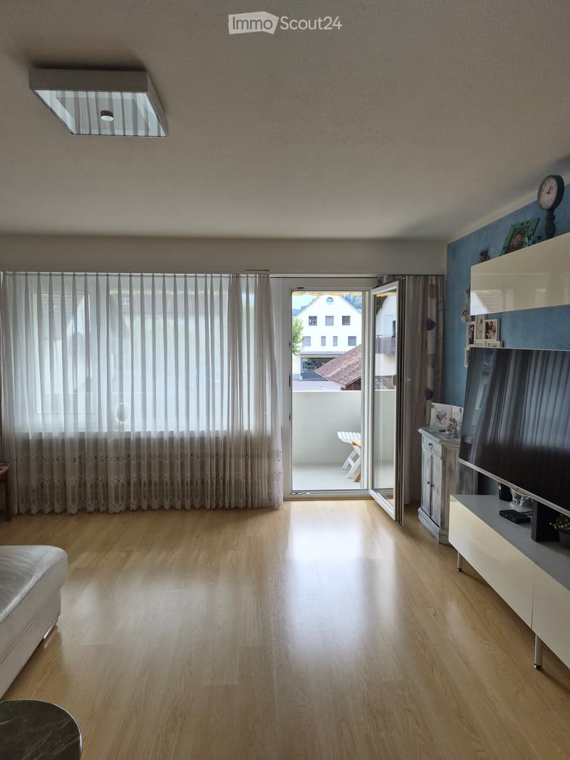 Bright living room with wooden flooring, large window with curtains, sliding glass door leading to a balcony, sofa, TV cabinet, and a small table.