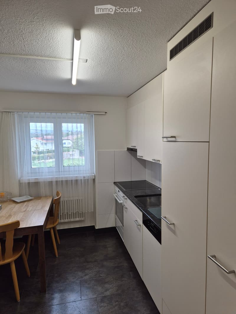 Modern kitchen with white cabinets, dark countertops, and a wooden dining table with chairs. Features an oven, refrigerator, and sink.