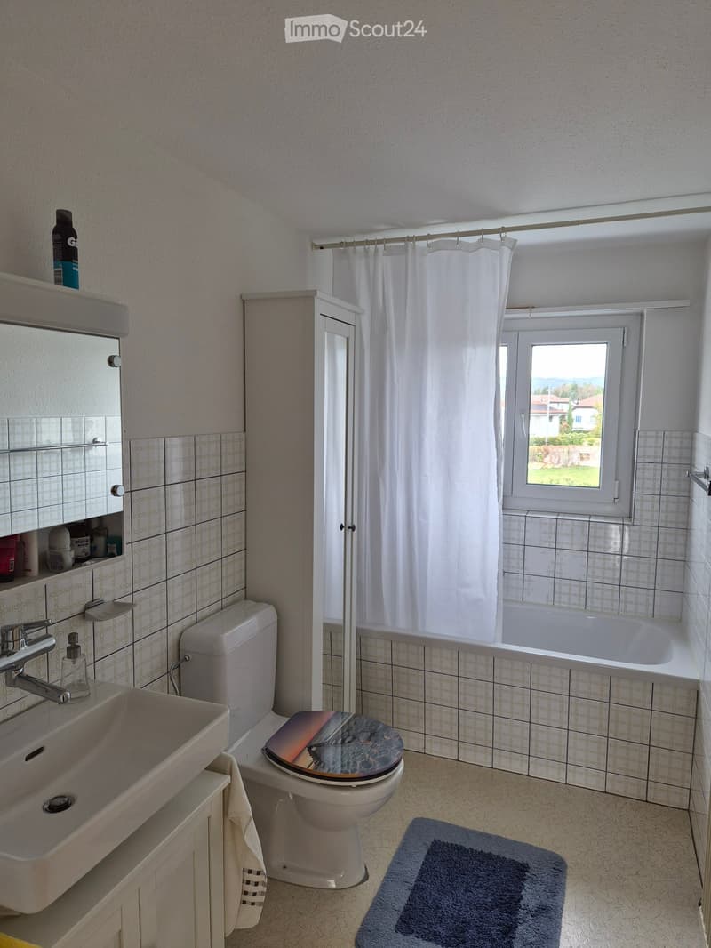 Bathroom with a sink, toilet, bathtub, mirror, and cabinet. A window with a view of the landscape is visible, along with a shower curtain and a blue bathroom rug on the floor.