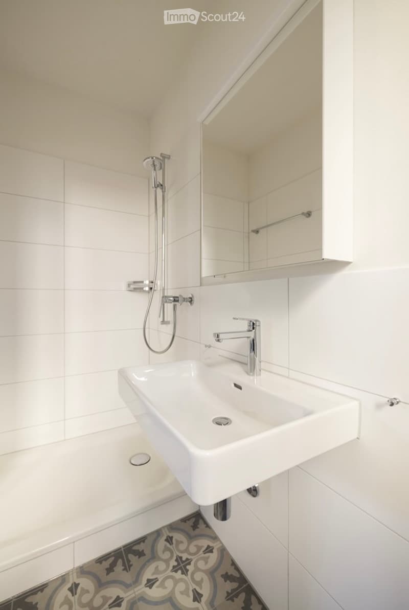 Modern bathroom with white tiles, a white sink, chrome faucet, and a showerhead. The floor features a decorative tile pattern.