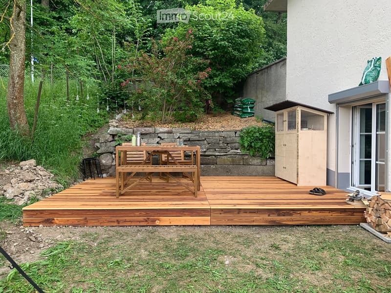 Wooden deck with a table, bench, and stool, surrounded by greenery and a stone wall. A small cabinet and shoes are visible near the house entrance.