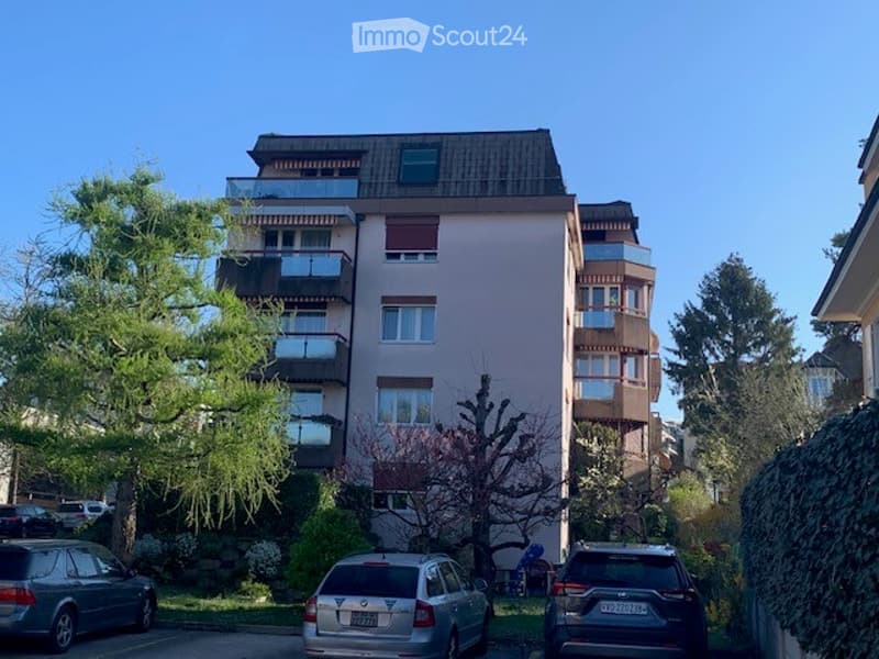 A multi-story residential building with balconies, surrounded by trees and parked cars.
