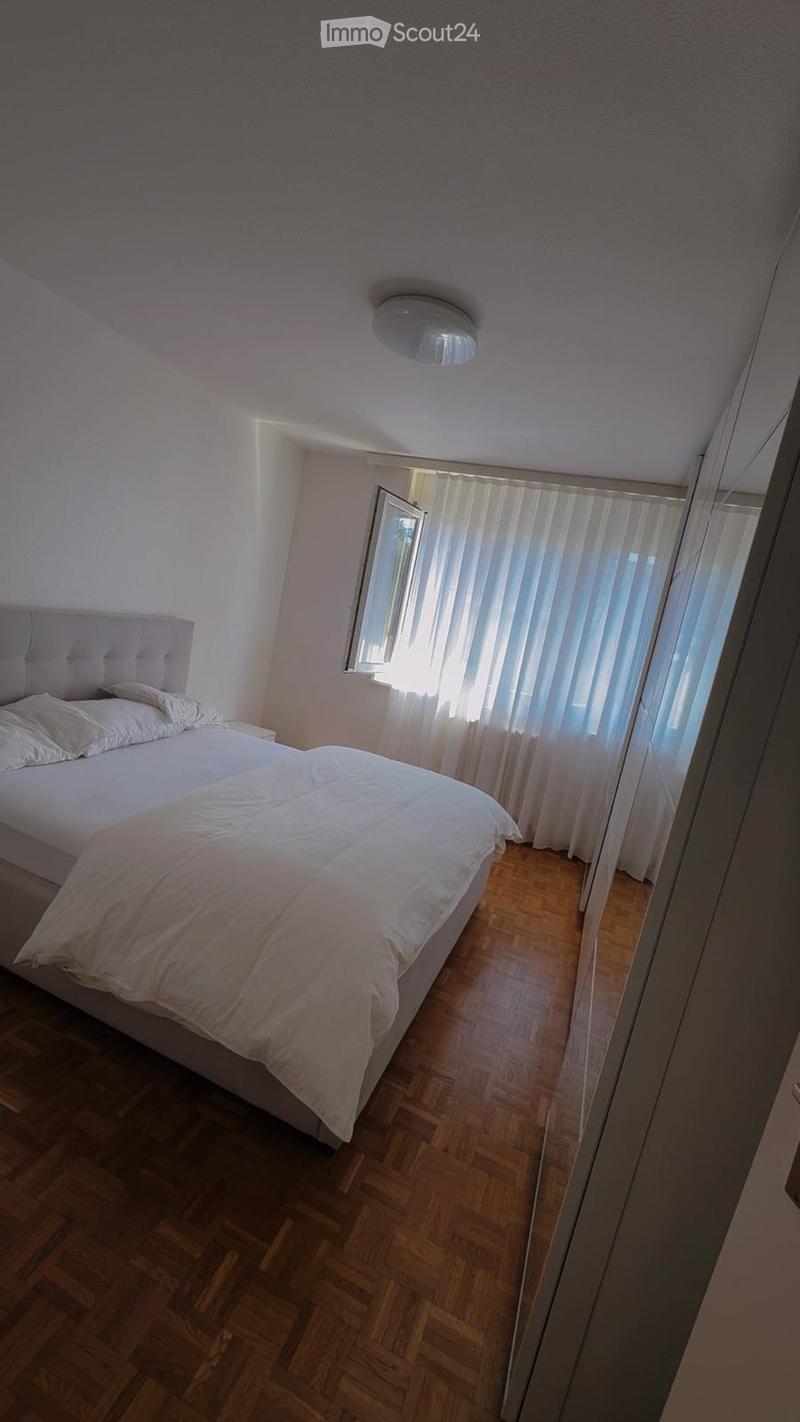 A modern bedroom with a large bed, white bedding, and wooden flooring. The room features a window with sheer curtains and a sleek, contemporary design.