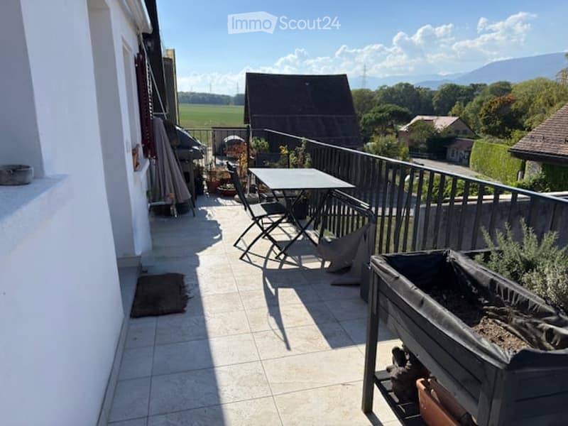 Balcony with tiled floor, metal railing, outdoor table and chairs, umbrella, and potted plants. Scenic countryside view in the background.