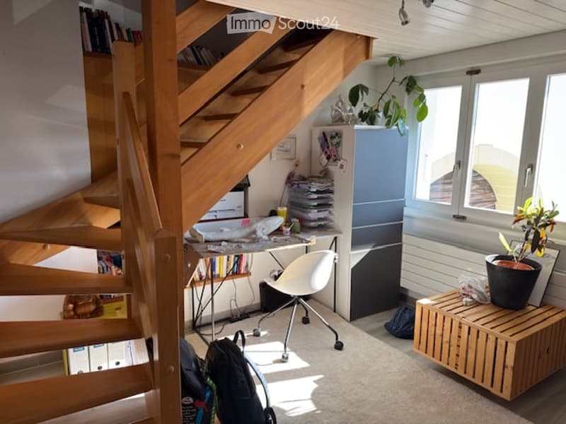 A modern, well-lit room with a wooden staircase leading to an upper level. The space includes a desk with a chair, a filing cabinet, a small wooden stool, and a potted plant on the windowsill. A backpack is placed on the floor near the desk.