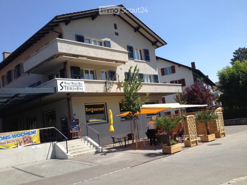 Two-story building with a restaurant sign, outdoor seating area, and a small garden with potted plants.