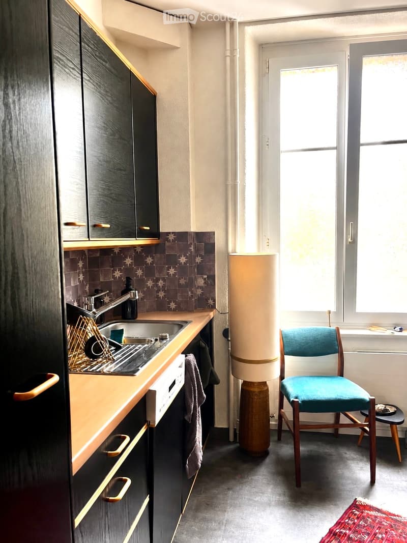 Modern kitchen with dark wooden cabinets, a countertop, sink, and a small seating area with a chair and stool. Features a decorative lamp and a patterned rug on the floor.