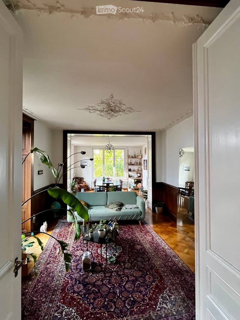 Living room with a teal sofa, coffee table, dining area with table and chairs, large window, and decorative plants.