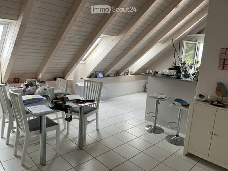 Bright, open-plan attic space with wooden slanted ceilings and large windows. Features a dining area with a table and chairs, a bar counter with stools, and a small cabinet with decorative items.