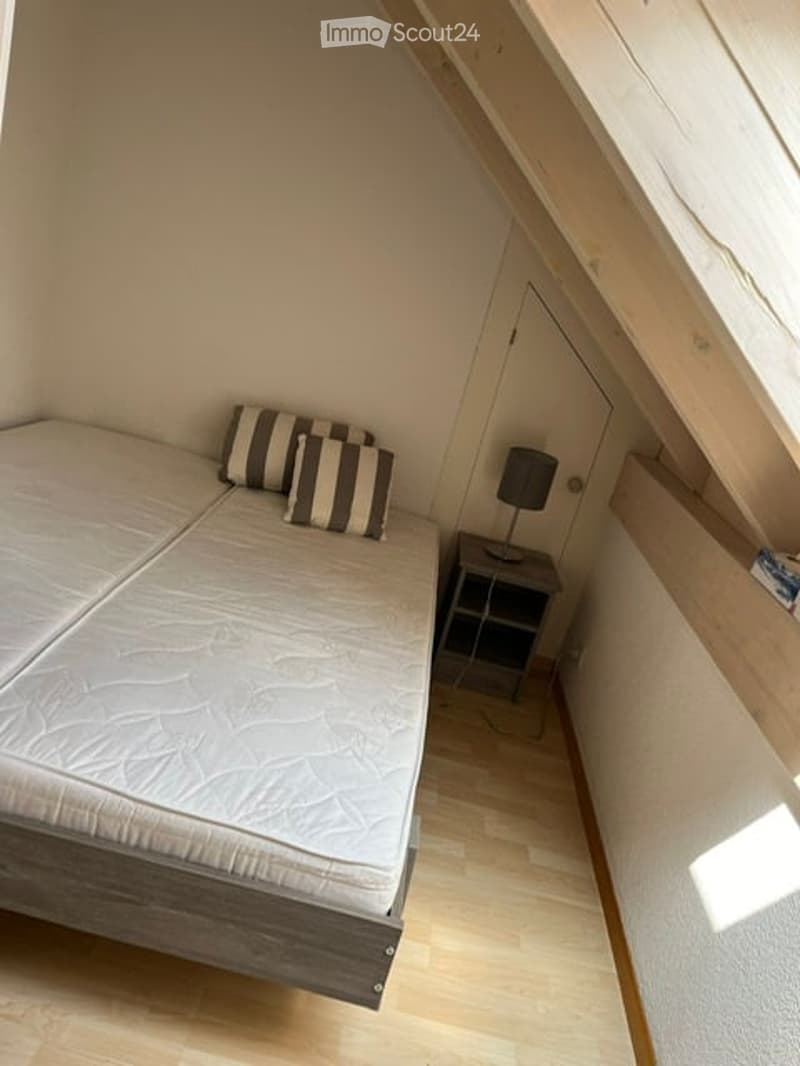 Cozy attic bedroom with a single bed, nightstand, lamp, and striped pillows. Light wooden flooring and slanted ceiling beams.