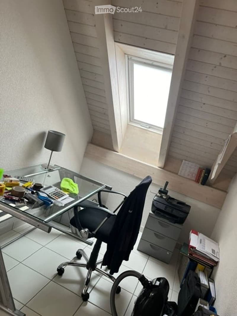A small, well-lit room with a slanted wooden ceiling and a skylight window. The room features a glass desk with a black office chair, a small lamp, and various office supplies. A printer sits on a gray cabinet, and books and a calendar are visible on a sh