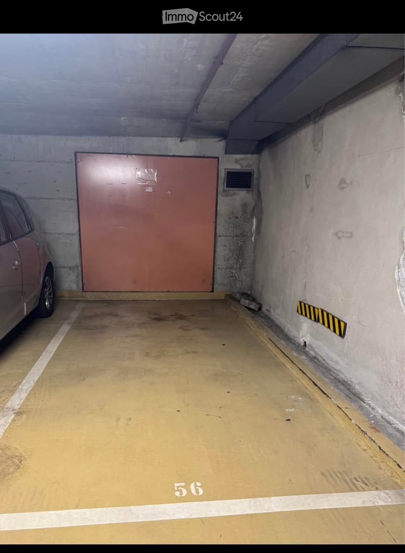 Underground parking space with concrete walls, a single car partially visible, a large red garage door, and a ventilation grate on the wall.