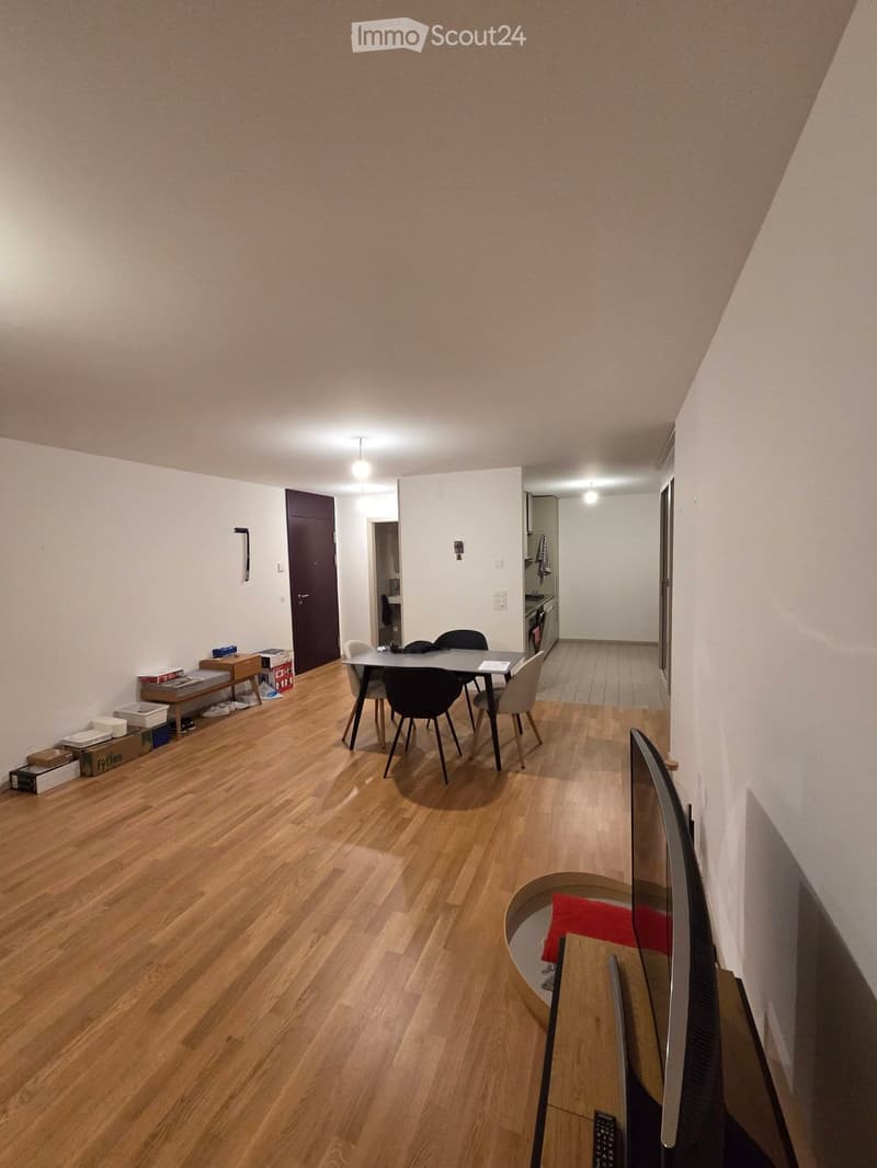 A furnished room with wooden flooring, a table with chairs, a TV screen, and a remote control on a small table. There are boxes and storage items near the door.