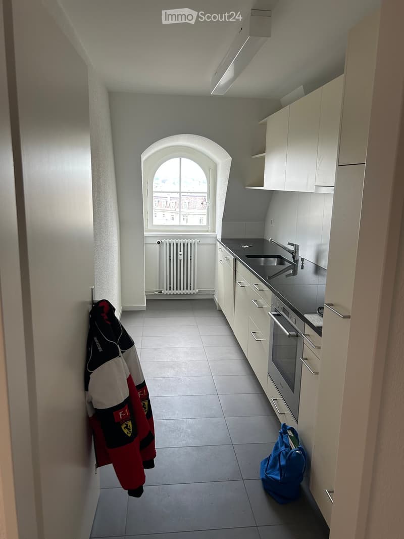 Small kitchen with white cabinets, a sink, a faucet, an oven, and a radiator. A jacket and a backpack are placed on the floor.