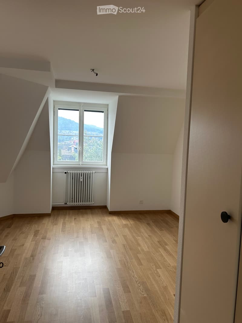 Empty room with wooden flooring, white walls, and a large window offering a scenic view of mountains and cityscape.