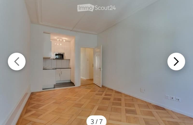 Empty room with light blue walls and wooden herringbone flooring. An open doorway leads to a small kitchen area with white cabinets and a microwave.
